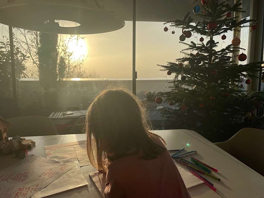 Girl from behind sitting at a table writing, Christmas tree in background and sunrise